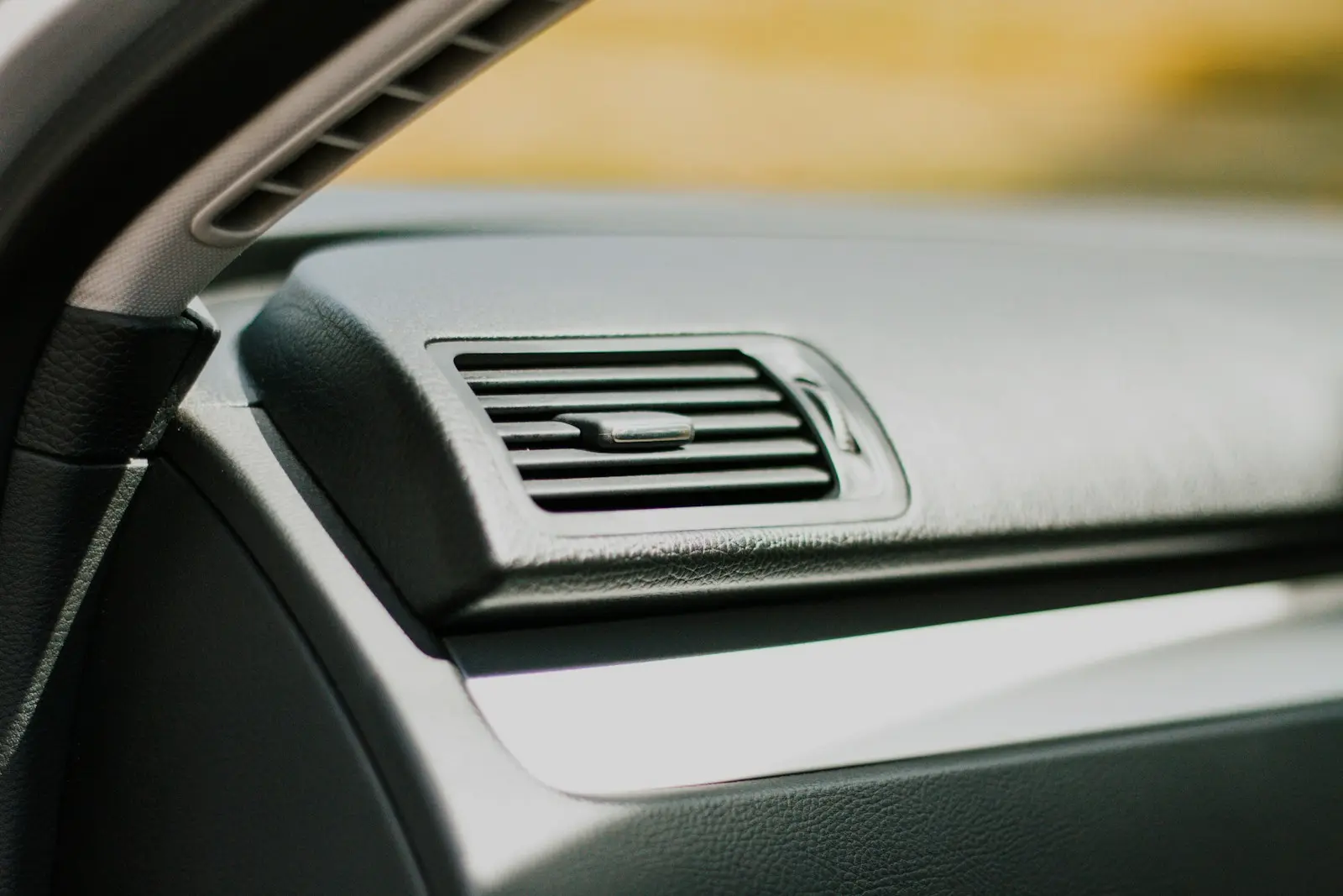Close-up of a rectangular dashboard air vent with horizontal louvres on a grey car dashboard, lit by daylight through the windshield.
