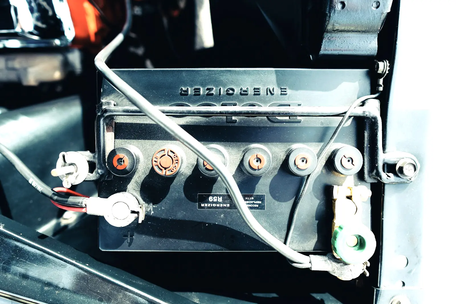 A car battery held down by a metal bracket, with cable clamps bolted to the positive and negative posts and red cell caps along the top.