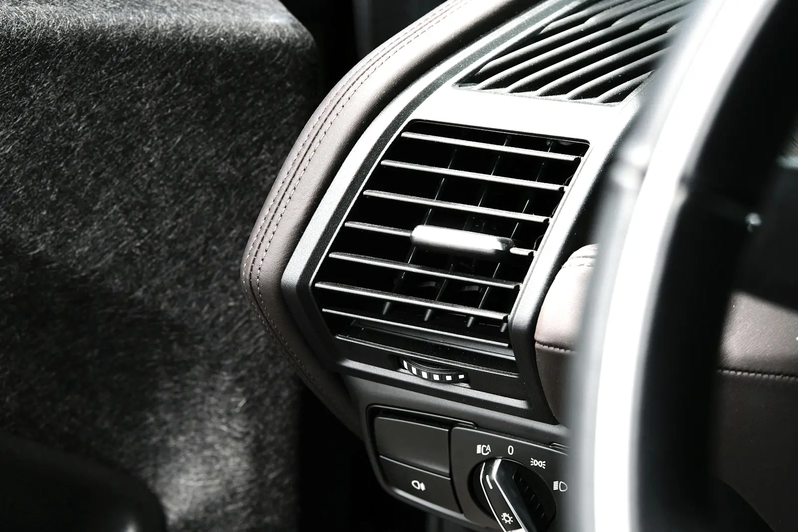 Close view of a dashboard air vent beside the steering wheel, with its louvres, airflow thumbwheel and the light switch below.