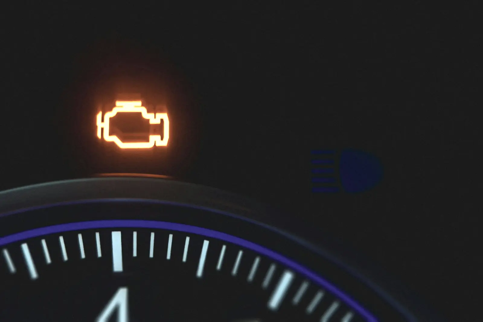 An amber engine-shaped check engine warning light glowing on a dark instrument cluster above the edge of a lit gauge.
