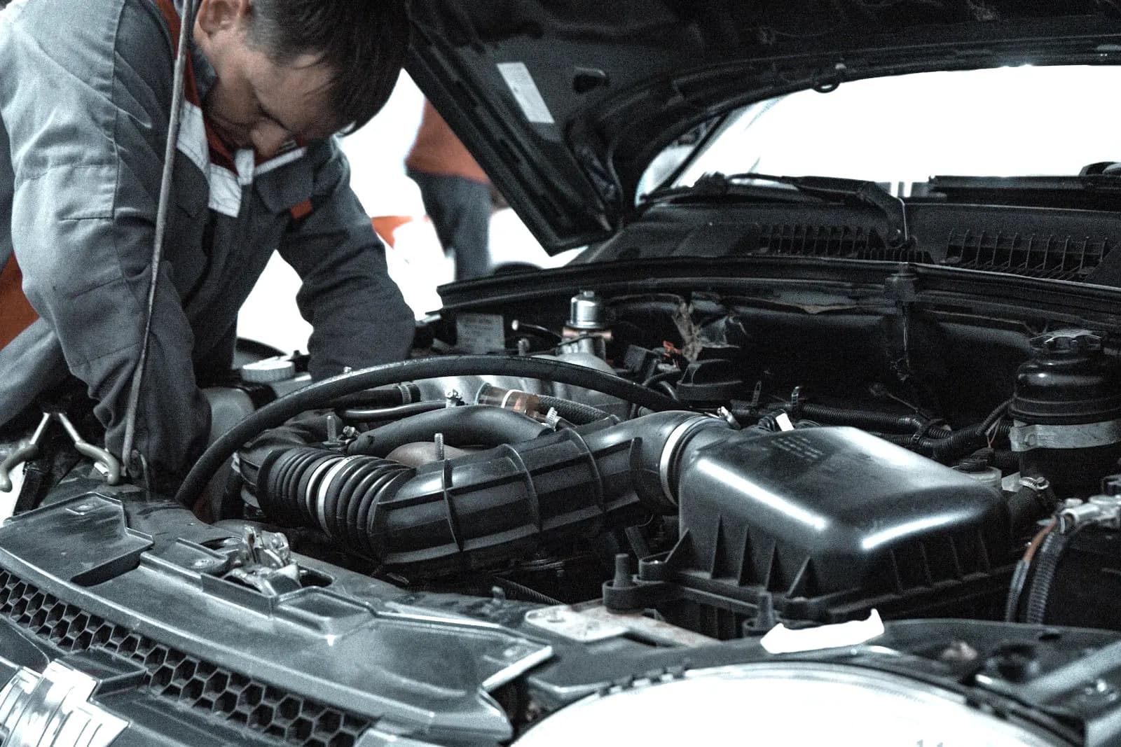 Open engine bay of a modern car with a mechanic leaning over it, showing the black plastic air filter box and the ribbed intake hose leading to the engine.
