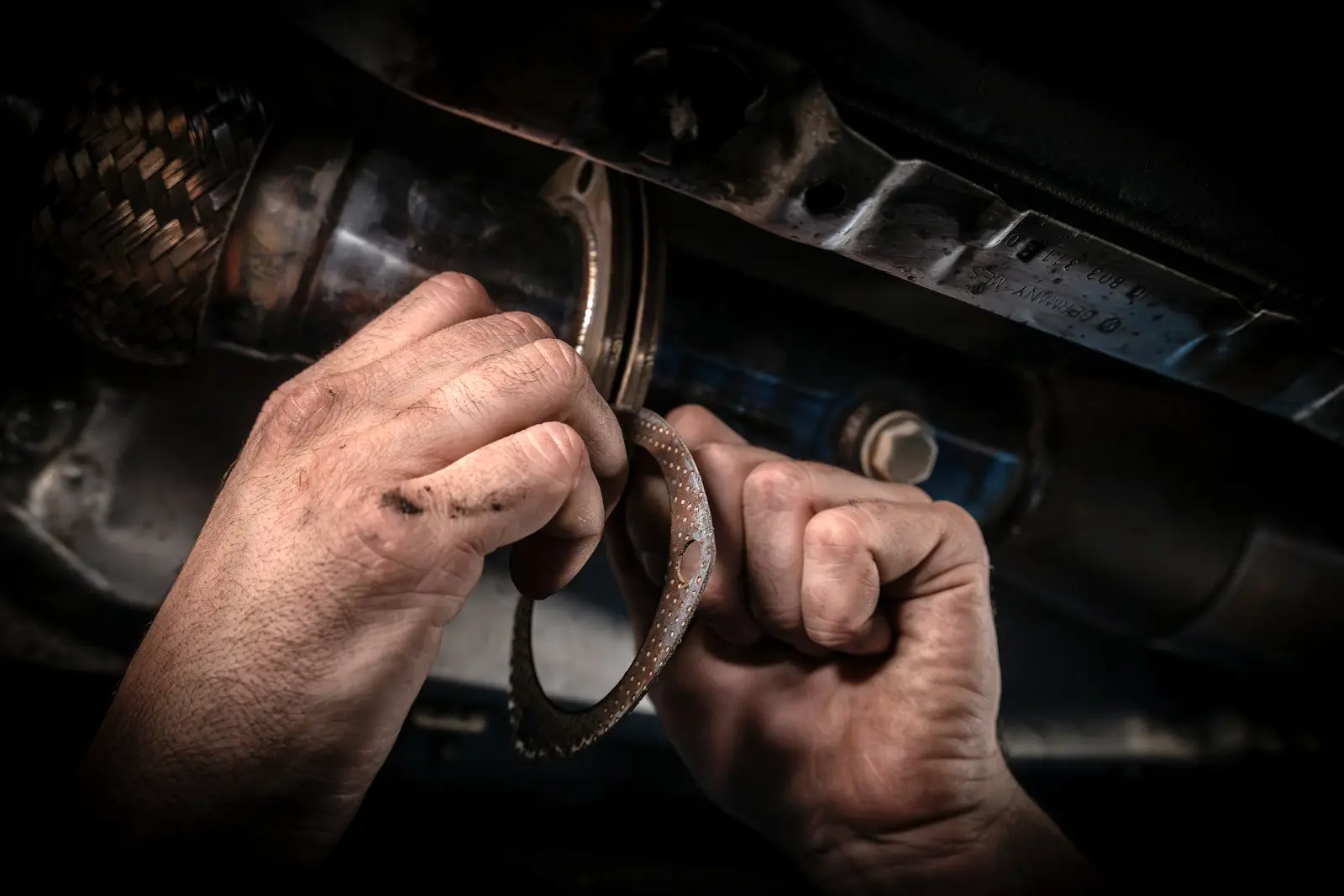 Two hands fitting a round exhaust gasket at a flanged joint under a car, next to a braided flex pipe section.