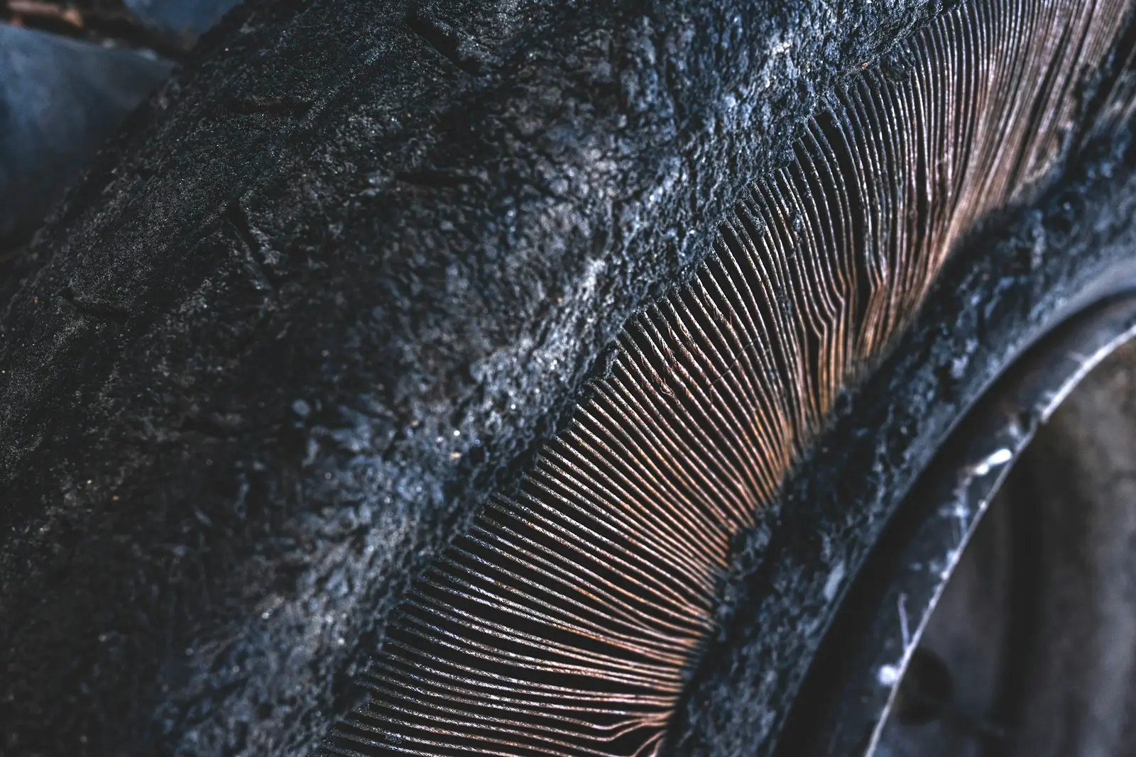 Close-up of a failed car tire where the rubber has split open along the sidewall, exposing rows of fine rusted cords underneath.