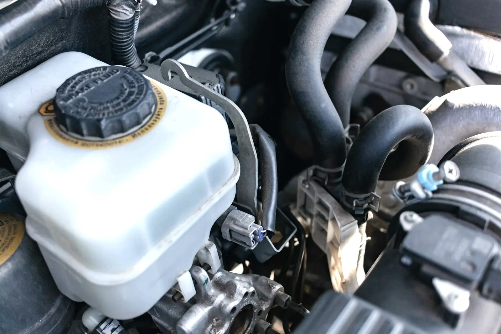 A translucent white brake fluid reservoir with a black screw cap, mounted on the master cylinder in a car engine bay, with hoses behind it.