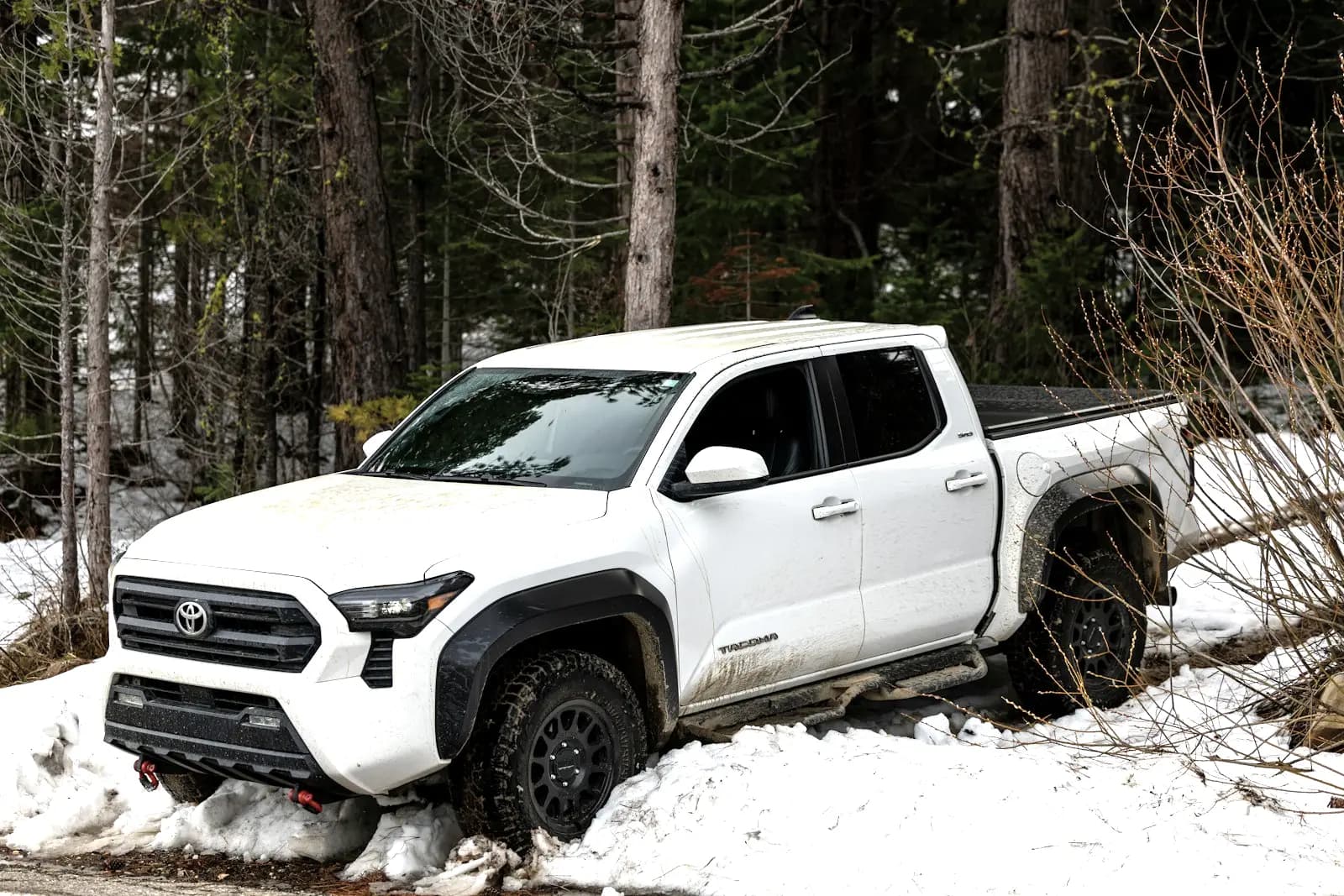 White pickup truck with mud on its doors, parked in deep snow at the edge of a forest track.