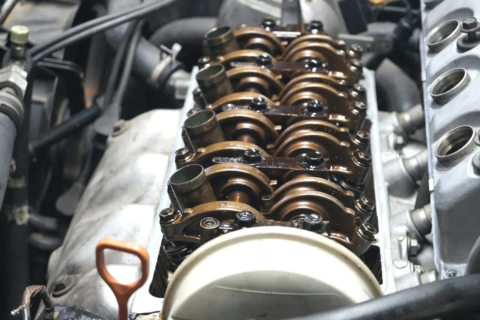 An engine with its valve cover removed, showing oil-coated rocker arms and camshaft, the cover's gasket sealing edge, and an orange dipstick handle.