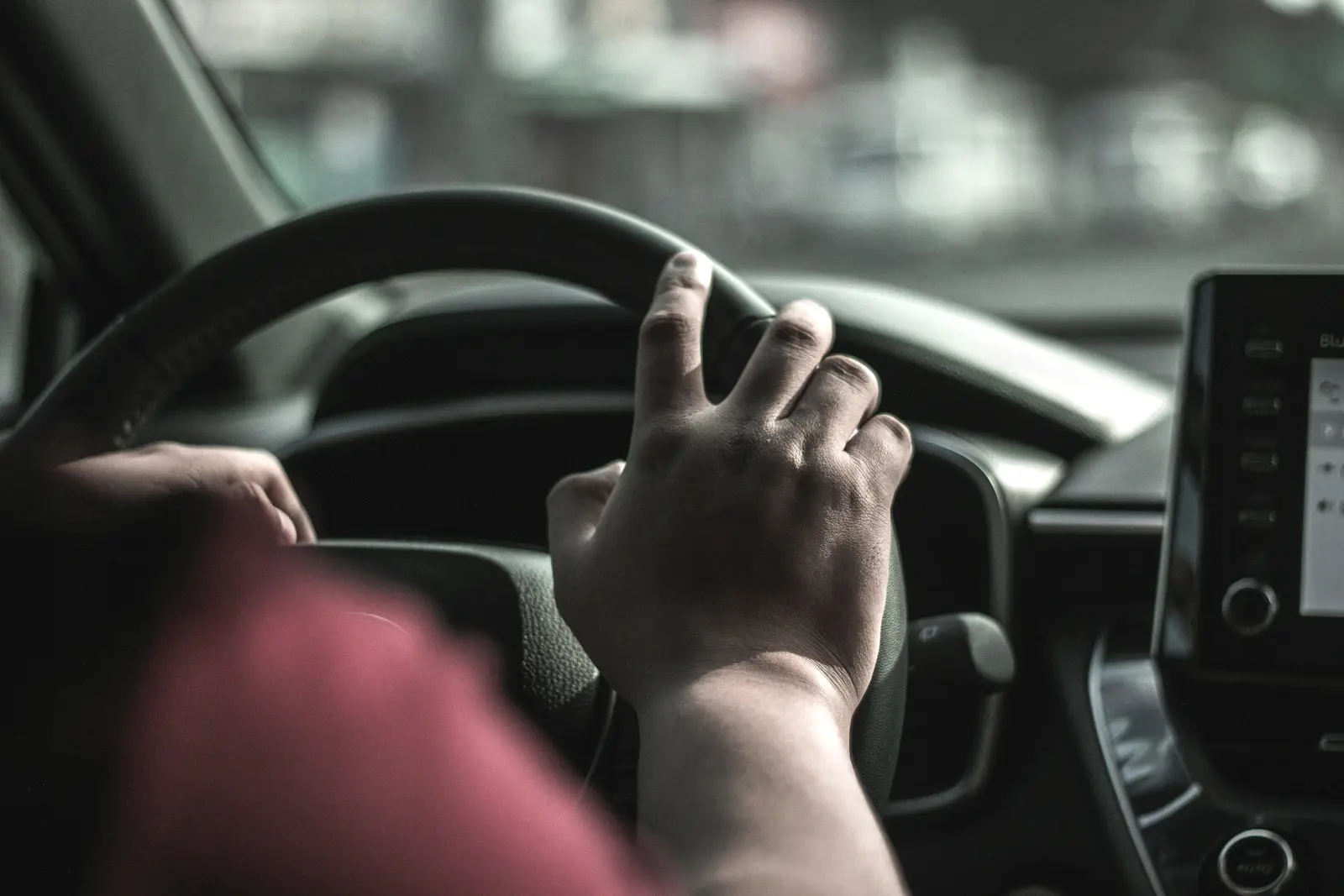 Driver's hand palming the top of the steering wheel through a turn, with the dashboard and centre screen behind it.