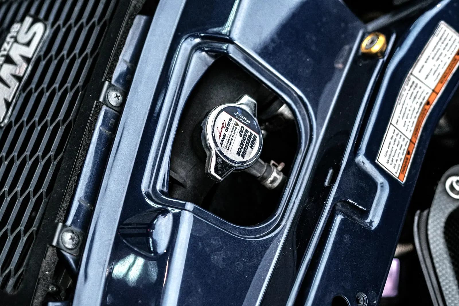 A chrome radiator cap stamped with its pressure rating, seen through an opening in a dark blue radiator support panel, with a warning label beside it.