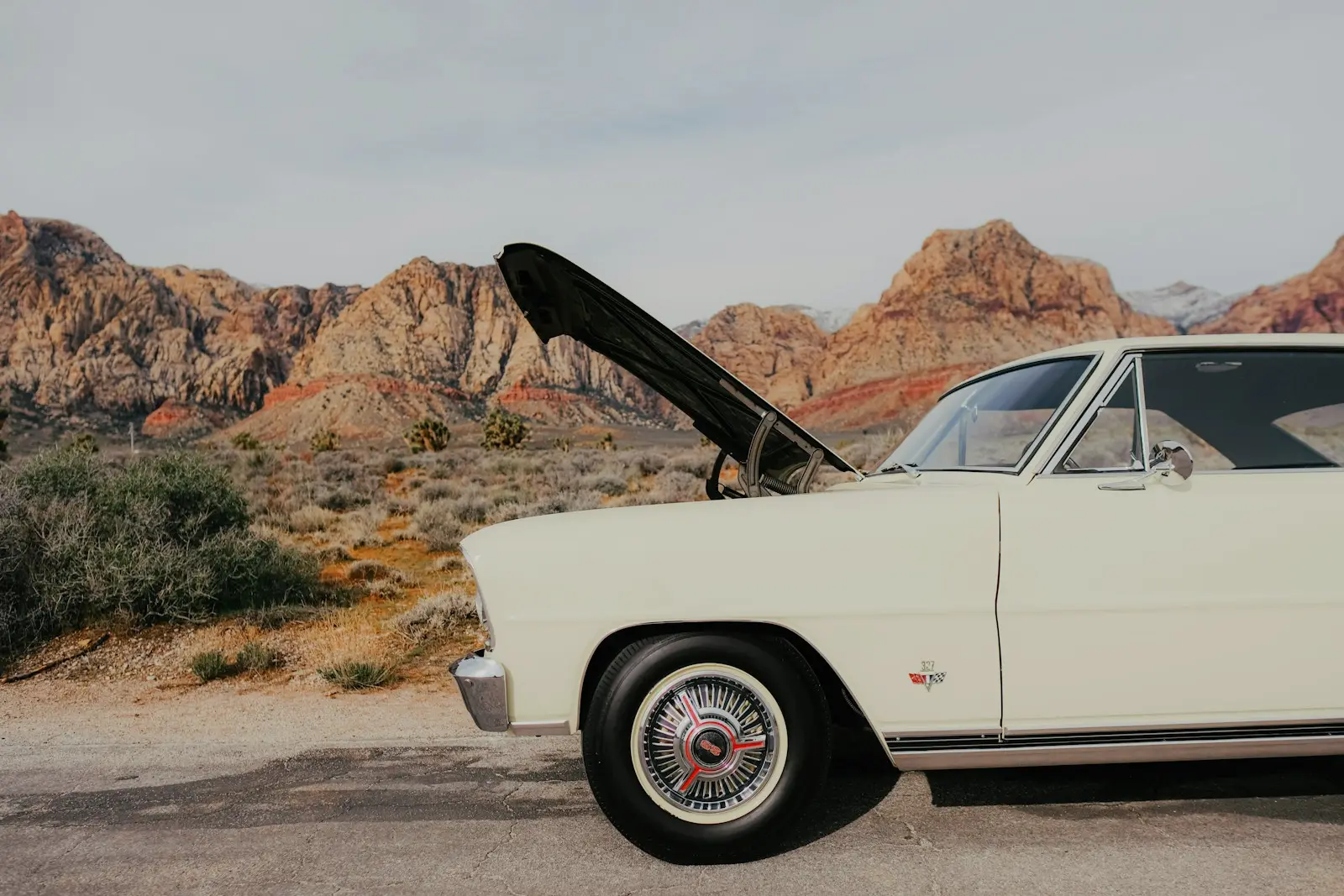 Cream classic car pulled off onto a gravel shoulder with its hood raised, desert scrub and red rock hills behind it.