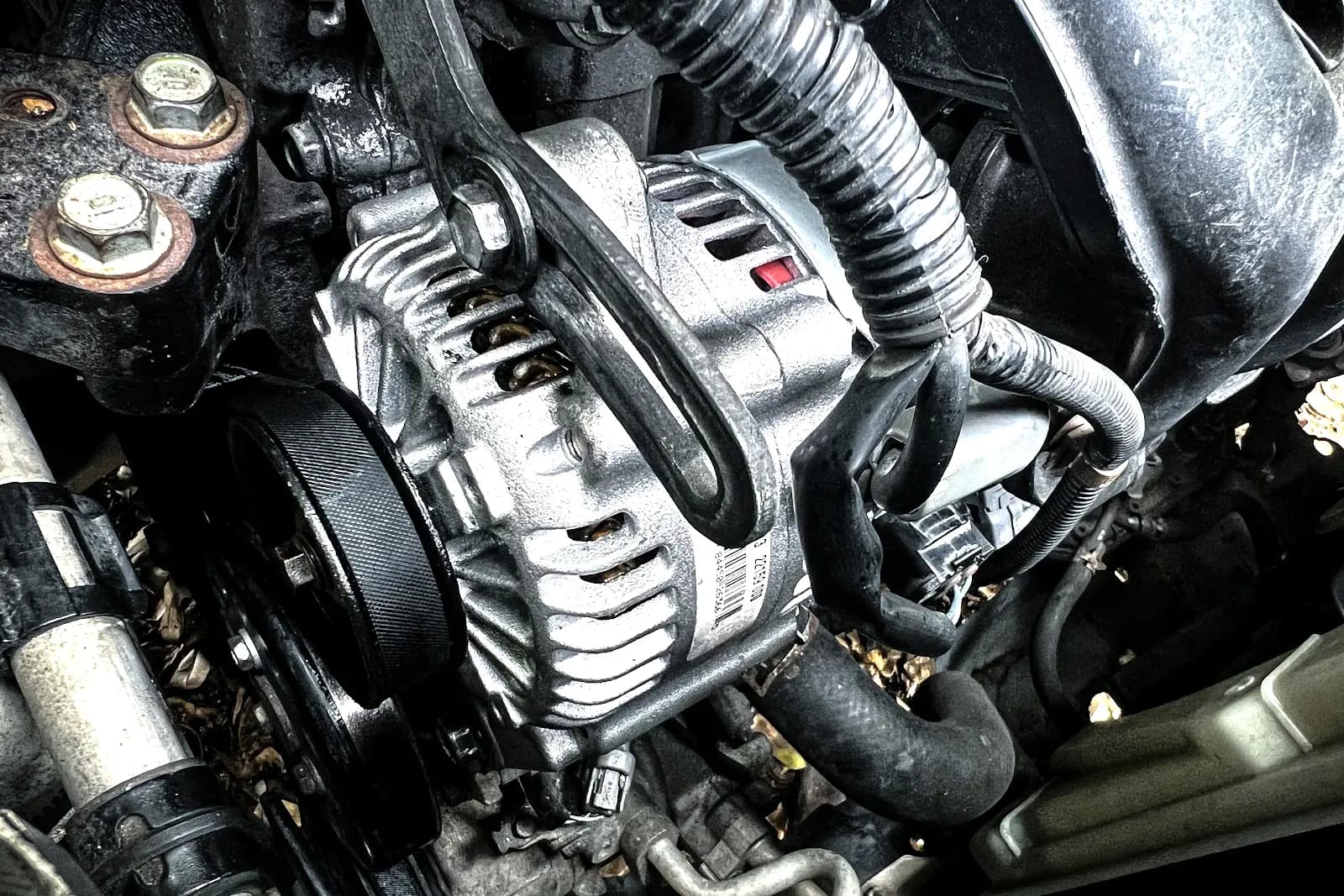Close-up of an alternator mounted on a car engine, with a ribbed drive belt wrapped around its pulley at the left.