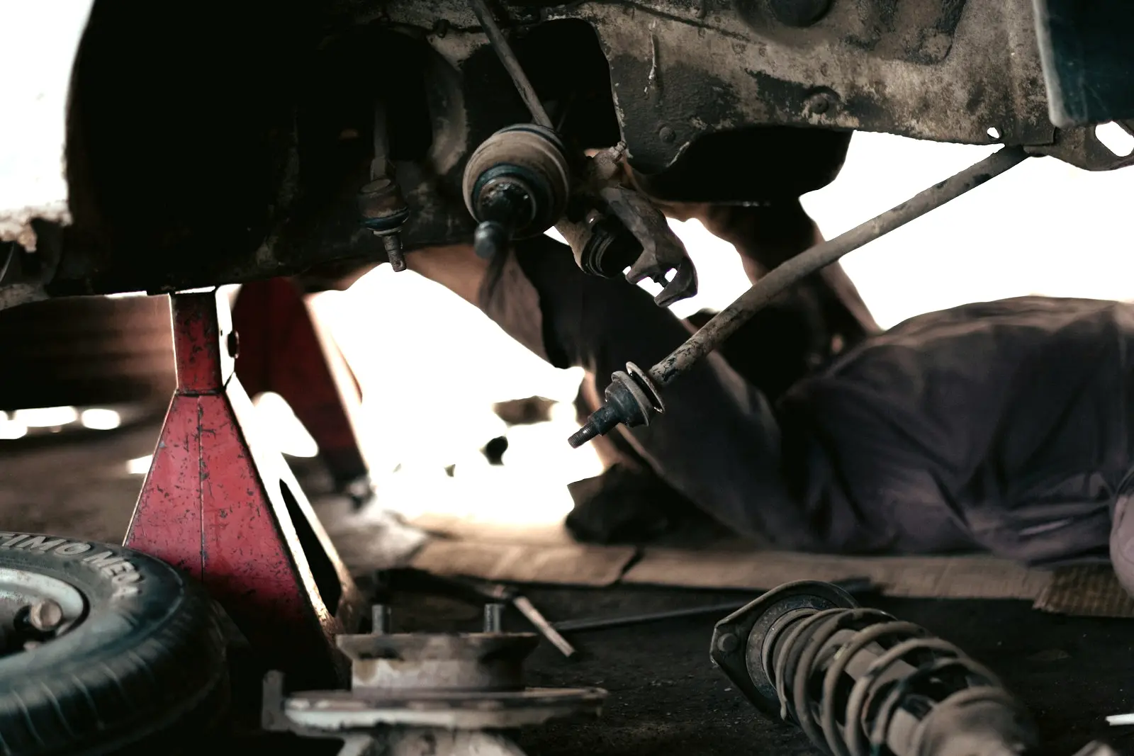 Car raised on a red jack stand with its front wheel off, and a removed strut with its coil spring lying on the floor.