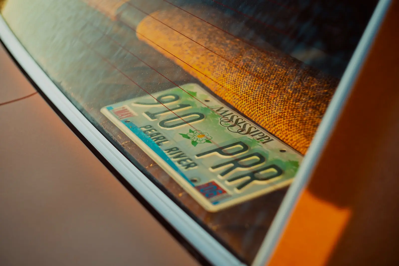 An old Mississippi license plate lying inside the rear window of a copper-colored car, lit by low evening sun.