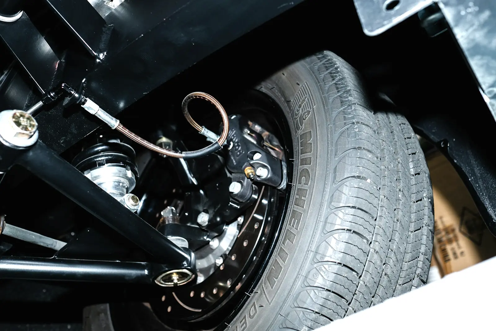 Wheel well seen from below, showing a black control arm with its pivot joints, the coil-over shock mount, brake caliper and tire.