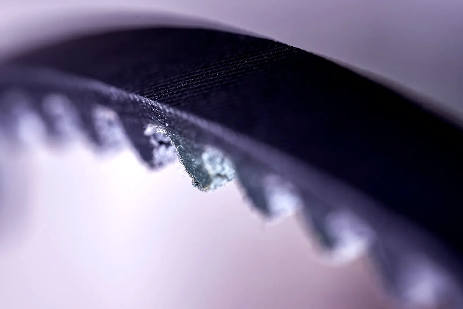 Extreme close-up of a black toothed rubber belt, with one tooth chewed and frayed, against a pale purple background.