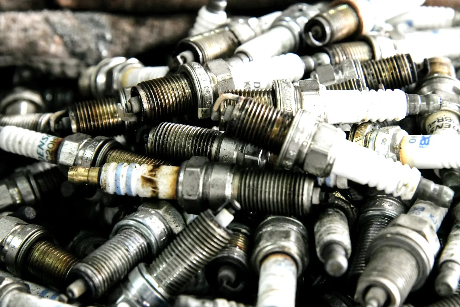 A pile of used spark plugs of several types, with white ceramic insulators and threaded ends stained brown and black from use.