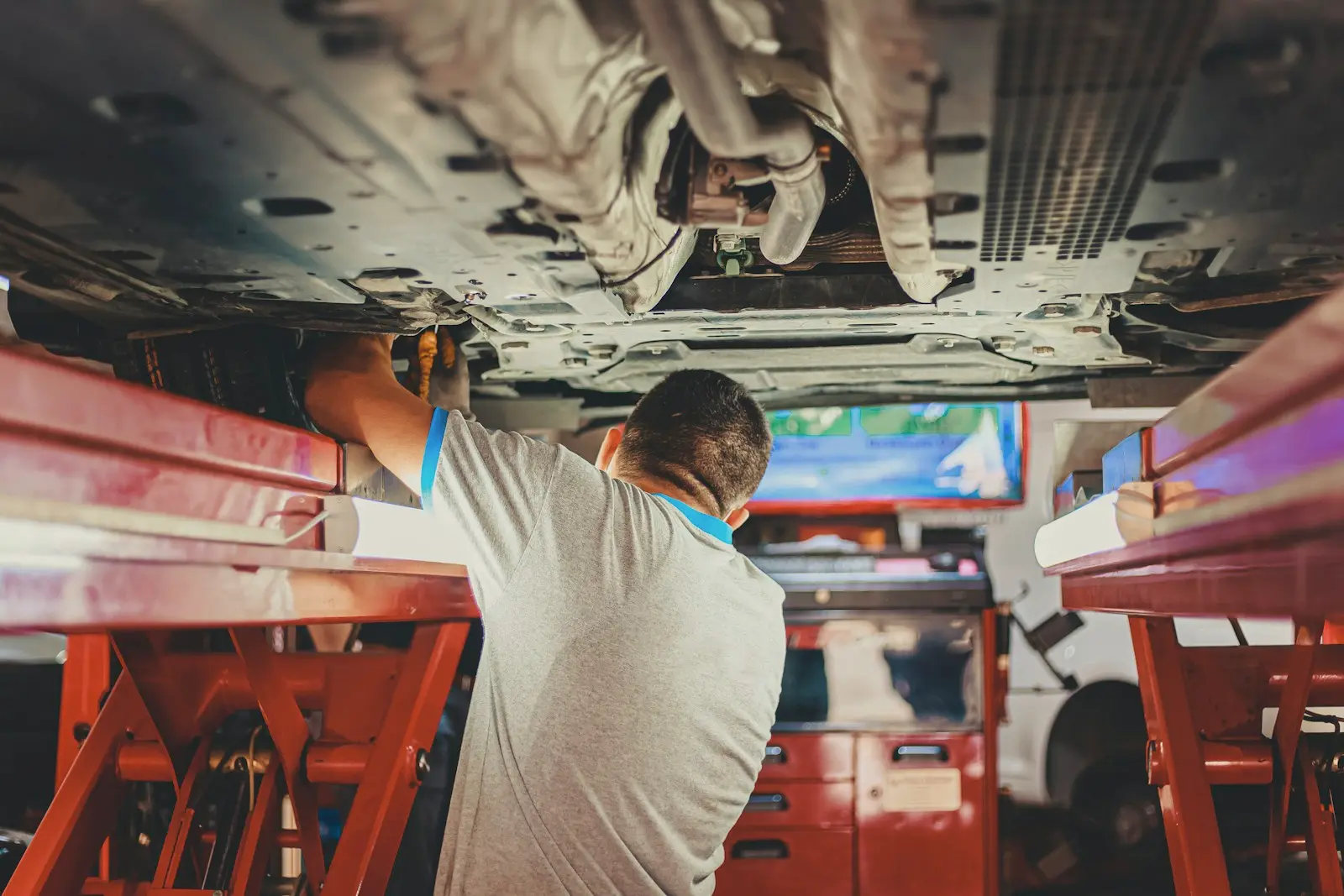 A mechanic seen from behind, reaching up to the underside of a car raised on a red scissor lift in a workshop.
