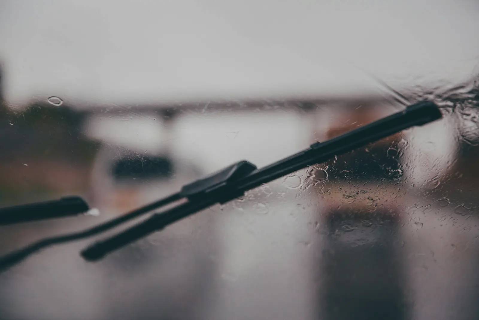 A black windshield wiper blade resting on rain-covered glass, with a blurred grey road and overpass visible through the windshield.