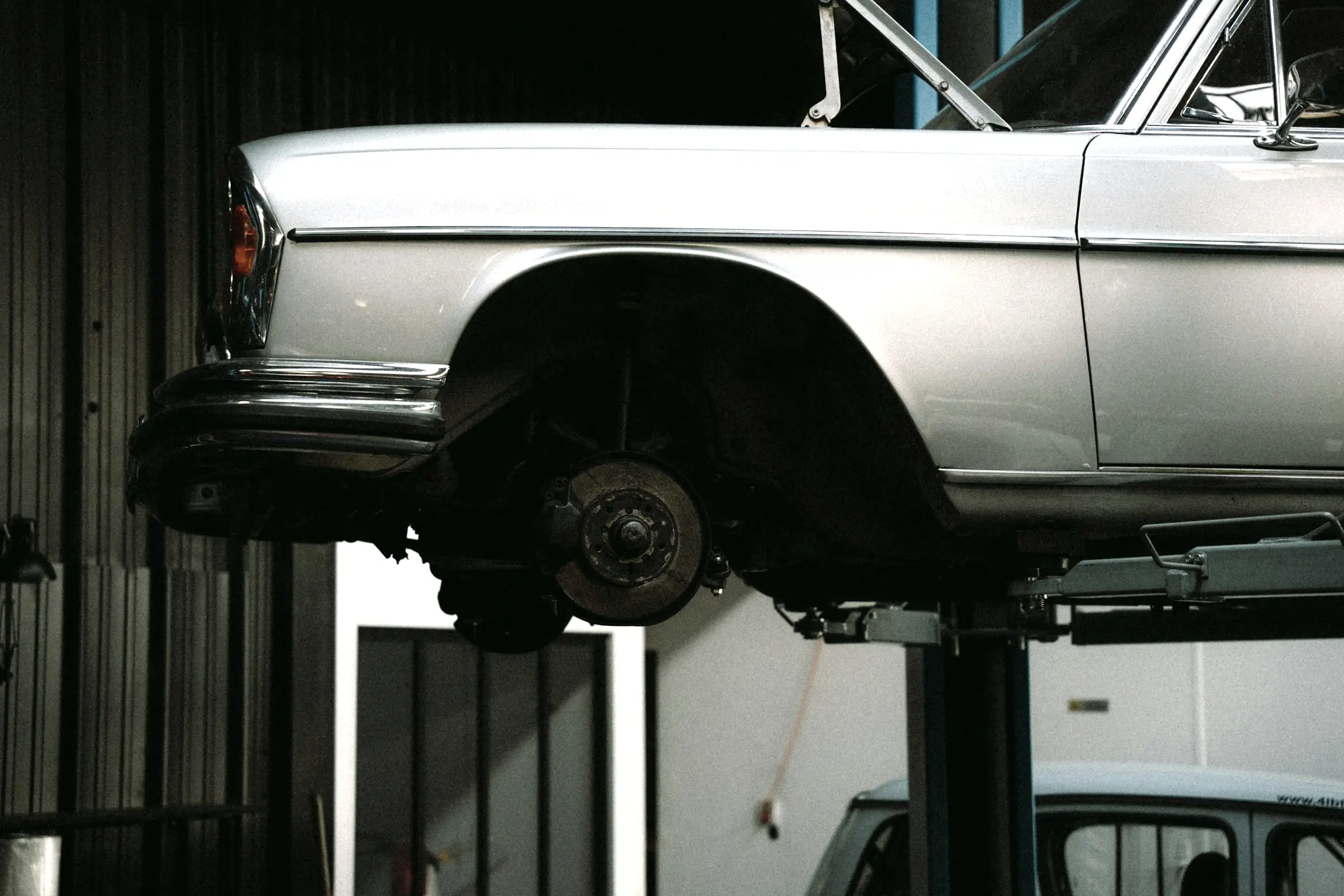 Silver sedan raised on a two-post lift in a workshop with its rear wheel removed, exposing the brake rotor, hub and suspension.